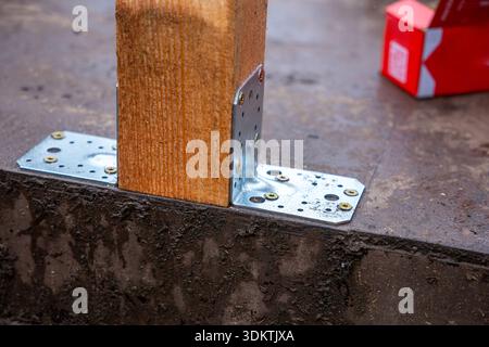 Holzpfosten, die mit Metallhalterungen und -Schrauben an einem Betonsockel befestigt sind, mit einer feuchten Oberfläche und einem roten Kasten im Hintergrund. Stockfoto