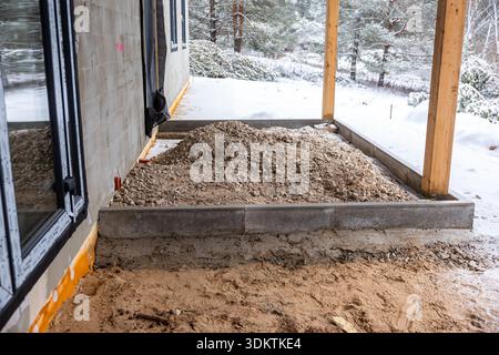 Baustelle im Freien mit rechteckigem Fundament, Kies, Holzpfosten und schneebedecktem Boden umgeben von Bäumen in winterlicher Umgebung. Stockfoto