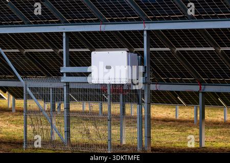 Reihen von Photovoltaikpaneelen auf einem Metallrahmen mit einem weißen Schaltkasten, der in einem umzäunten Bereich auf trockenem Gras mit sichtbaren roten Bändern untergebracht ist. Stockfoto