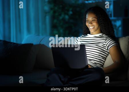 Glückliche schwarze Frau, die ihren Laptop überprüft und nachts zu Hause auf einer Couch sitzt Stockfoto