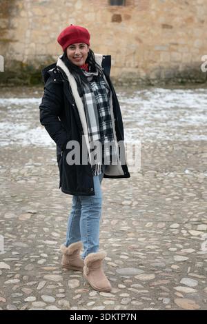 Junge Erwachsene Frau mit Wintermantel, roter Baskenmütze, kariertem Schal, Jeans und Stiefeln, draußen stehen Stockfoto