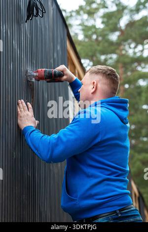 Ein Mann in einem blauen Kapuzenpullover bedient ein Elektrowerkzeug an einer dunklen, vertikal verglasten Wand eines Hauses, das im Bau ist und von hohen Bäumen und Materialien umgeben ist. Stockfoto