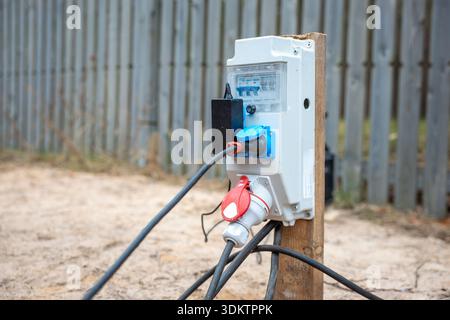 Elektrischer Verteilerkasten im Freien an Holzpfosten mit transparenter Abdeckung, Leistungsschalter, roten und blauen Steckern und Kabeln auf der Baustelle. Stockfoto