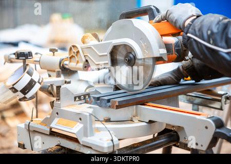 Nahaufnahme einer Gehrungssäge in Gebrauch, mit Handschuhen, die Holz durch die Klinge führen. Sägemehl und verschwommene Baumaterialien füllen den Hintergrund. Stockfoto