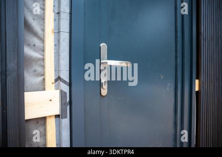 Nahaufnahme einer modernen Tür mit Metallgriff in dunkelblauem Rahmen, umgeben von Holzbalken und Isolierstoff auf einer Baustelle. Stockfoto