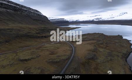 Aus der Vogelperspektive schlängelt sich eine gewundene Straße durch die zerklüftete, schneebedeckte Landschaft, im Kontrast zum ruhigen, reflektierenden Wasser unter einem glühenden Himmel, S Stockfoto