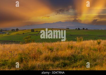 Goldene Sonnenstrahlen, die durch einen lebendigen Sonnenuntergangshimmel über sanfte grüne Felder und ferne Berge Streifen Stockfoto