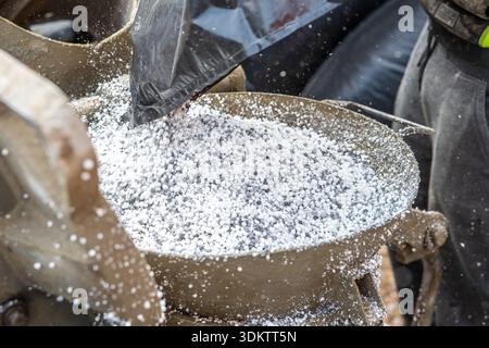 Nahaufnahme eines Metallbehälters, da weißes Granulat aus einem schwarzen Beutel gegossen wird, wobei die Maschine und ein Arbeiter an der Seite teilweise sichtbar sind. Stockfoto