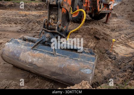 Nahaufnahme eines Baggerlöffels, der Erde auf einer Baustelle gräbt, mit Reifenspuren, loser Erde, einem gelben Kabel und einer orangefarbenen Übersichtsmarke Stockfoto