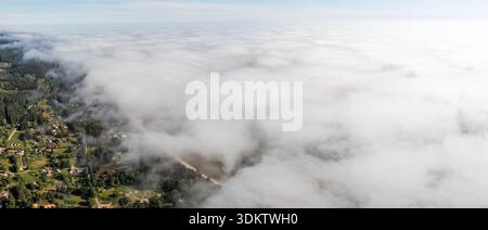 Aus der Vogelperspektive auf eine ländliche Gegend mit verstreuten Häusern, grünen Feldern und einem Wald, teilweise verdeckt von tief liegenden Wolken, die die Szene dominieren. Stockfoto