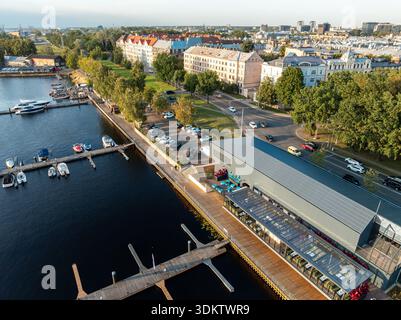 Eine Marina am Wasser mit kleinen Booten an Holzpiers, ein modernes Glasfassadengebäude mit Sitzgelegenheiten im Freien und ein farbenfrohes Gebäude im historischen Stil Stockfoto