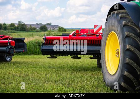 Nahaufnahme eines roten Mähaufbereiters an einem grünen Traktor mit großem gelbem Felgenrad auf Schnittweide. Zinken- und Aufbereitertrommeln zeigen Heu Stockfoto