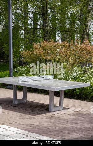 Tischtennisplatte im Freien mit einem Metallnetz auf gepflastertem Boden, umgeben von Büschen, blühenden Pflanzen, hohen Bäumen und einer Metallstange auf der linken Seite. Stockfoto