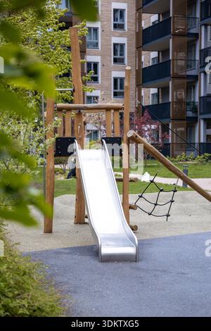 Moderner Spielplatz mit Metallrutsche, Holzrahmen und Kletternetz, umgeben von Wegen, Grün, und mehrstöckige Wohngebäude. Stockfoto