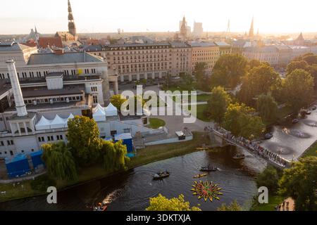 Das Denkmal der Freiheit in Riga, Lettland, steht in der Nähe eines Kanals mit Booten und einer schwimmenden Installation, umgeben von Bäumen, Wegen und historischen Gebäuden Stockfoto