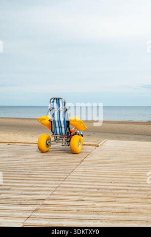Ein gestreifter blau-weißer Strandrollstuhl mit gelben Rädern und Armlehnen befindet sich auf einer Holzpromenade, die zu einem Sandstrand und dem ruhigen Meer führt. Stockfoto
