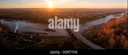 Die Luftaufnahme zeigt Kuldiga in Lettland bei Sonnenuntergang, die alte Ziegelbrücke über die Venta, Venta Rapid auf der linken Seite, Herbstbäume, Kirchtürme, warmes Licht und Stockfoto