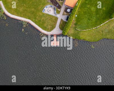 Aus der Vogelperspektive sehen Sie ein Dock mit einem roten Dach, das sich ins Wasser erstreckt, einen gekrümmten Steinweg, einen runden Sitzbereich und ein rot gekacheltes Dachgebäude. Stockfoto