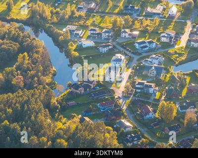 Aus der Vogelperspektive eines Wohnviertels mit gewundenen Straßen, einem reflektierenden Wasserweg, offenen Grünflächen und Baumgruppen in sanftem Sonnenlicht. Stockfoto