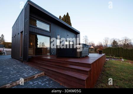 Moderne Kabine mit schrägem Dach und vertikaler schwarzer Verkleidung auf einer breiten Holzterrassenterrasse mit großem Whirlpool, Glaspaneele reflektieren spätes d Stockfoto
