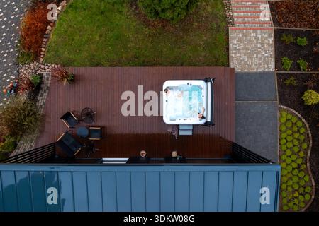 Zwei Personen entspannen sich in einem quadratischen Whirlpool auf einer Holzterrasse neben einem Metalldach, mit Terrassenstühlen und einem runden Tisch. Die Luftaufnahme hebt starke Linien und hervor Stockfoto