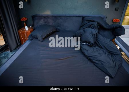 Modernes Schlafzimmer mit dunkler strukturierter Wand, schwarzer Bettwäsche, Nachttischen aus Holz mit orangefarbenem Blumendekor und Tageslicht durch große Fenster. Stockfoto