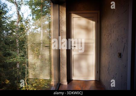 Moderner Innenraum mit weißer Holztür an einer strukturierten Wand. Sonnenlicht strömt durch ein großes Glasfenster und offenbart hohe grüne Bäume Stockfoto