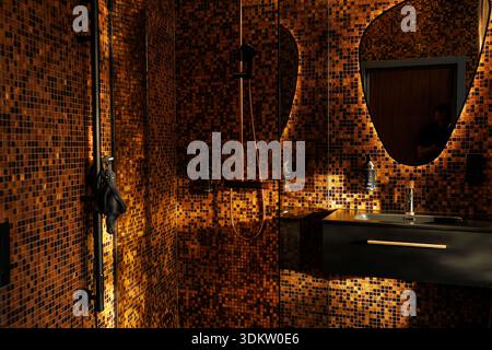 Modernes Bad mit warmen orangefarbenen und braunen Mosaikfliesen, Wandwaschbecken mit Beleuchtung, Regendusche und einzigartig geformten Spiegeln Stockfoto