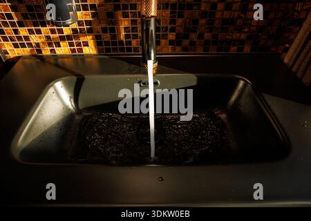 Ein schwarzes Waschbecken mit Wasser aus einem metallischen Wasserhahn mit kupferfarbenem Griff, das an einer Wand mit Mosaikfliesen in warmen Braun- und Goldtönen steht. Stockfoto