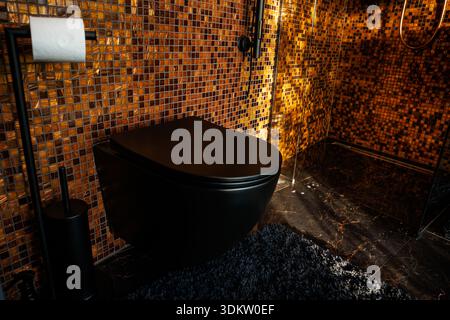 Modernes Bad mit orangefarbenen und goldenen Mosaikfliesen, schwarzer Toilette und Armaturen, dunklem Marmorfußboden und Glasdusche. Stockfoto