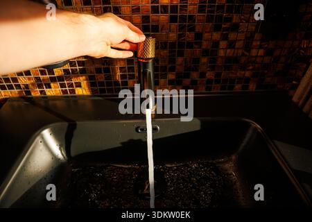 Eine Hand dreht sich an einem schwarzen Wasserhahn mit einem goldenen Griff, und lässt Wasser in ein dunkles Waschbecken frei. Warme Mosaikfliesen in Braun- und Bernsteintönen bilden die Kulisse. Stockfoto