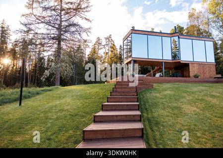 Modernes Holzhaus mit eckigem Design und großen Glaspaneelen auf einem grasbewachsenen Hang, umgeben von hohen Bäumen mit Sonnenlicht, das durch Äste filtert. Stockfoto