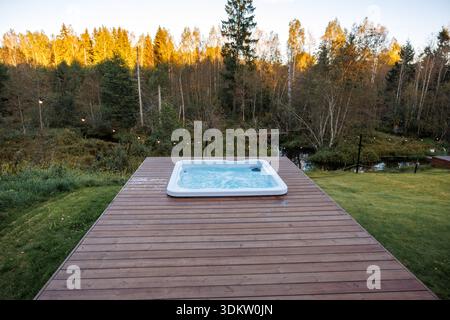 Eine Holzterrasse mit Whirlpool und Lichterketten befindet sich in einem Außenbereich mit Herbstbäumen. Die Terrasse überblickt ein kleines Wasserspiel bei Sonnenuntergang. Stockfoto