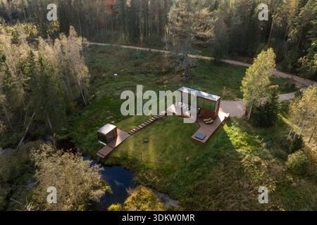 Moderne Kabine mit großen Glasfenstern, Holzterrasse und Sitzgelegenheiten im Freien. Eine kleinere Einheit, ein Holzweg und ein Bach sind von Bäumen umgeben. Stockfoto
