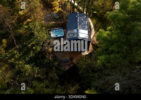 Eine moderne Hütte mit großen Glasfenstern, eckigem Dach und Holzterrasse mit Sitzgelegenheiten und Whirlpool, umgeben von Bäumen und Sonnenlicht, das nach außen filtert Stockfoto