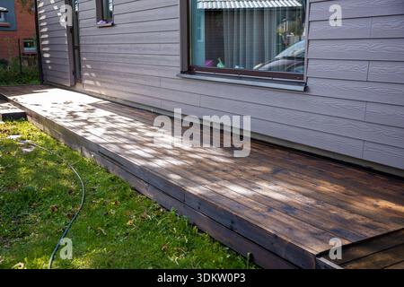 Eine Holzterrasse mit frischen Flecken, die an einem Haus mit hellgrauen Abschlüssen befestigt ist. Sonnenlicht zieht durch Bäume und ein Gartenschlauch liegt auf dem Gras in der Nähe. Stockfoto