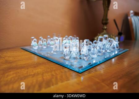 Die Schachfiguren aus Glas ruhen auf einem mattierten Glasbrett mit einer grünen Kante auf poliertem Holz. Niedriger Winkel, flacher Fokus Rahmen mit Kingsize- und Queensize-Bett, mit Messinglampe Stockfoto