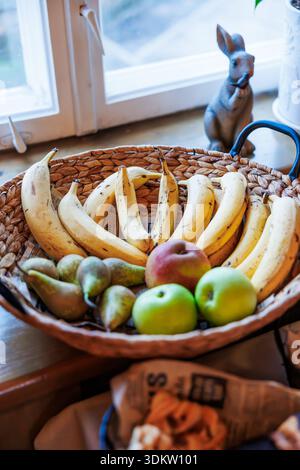 Ein gewebtes Tablett hält Bananen in einem Ring um grüne Äpfel, Birnen und einen Pfirsich auf einer Holzfläche am Fenster, mit einer kleinen Kaninchenfigur und einem weichen da Stockfoto