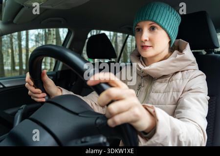 Eine Erwachsene Frau in einer Jacke sitzt hinter dem Steuer eines Autos. Die Frau ist die Fahrerin. Winter, ein Park vor dem Fenster Stockfoto
