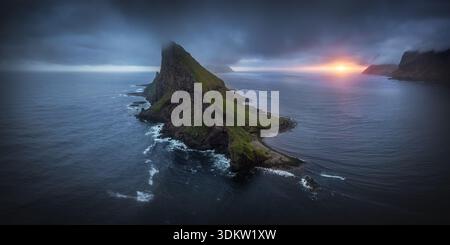 Aus der Vogelperspektive auf die Insel Tindhólmur, die in den stimmungsvollen Himmel hineinragt, mit ihren zerklüfteten Gipfeln und dem glatten, dunklen Meer, Bøur, Färöer-Inseln. Stockfoto