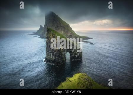 Aus der Vogelperspektive auf den dramatischen Bogen von Drangarnir und Tindhólmur, der aus dem stimmungsvollen Atlantik aufsteigt, wo dunkles Wasser auf grüne Klippen trifft, Sørvágur, Färöer I. Stockfoto