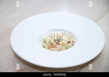 Gourmet-Risotto mit Reis, Speckbröseln, Parmesanspänen und frischen Kräutern, kunstvoll auf einer weißen, strukturierten Schüssel angeordnet, symbolisiert gehobene Küche und Stockfoto
