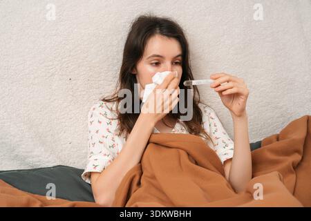 Eine junge Frau, die mit einer braunen Decke bedeckt ist, sitzt im Bett, hält ein Taschentuch an der Nase und blickt auf ein Thermometer. Konzept der Grippe, Fieber, Erkältung, Erkältung Stockfoto