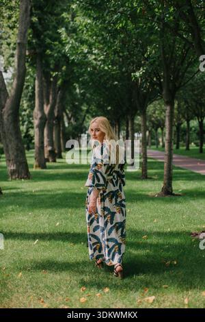 Frau, die in einem grünen Park in einem Blumenmaxi-Kleid läuft, Wind weht durch die Haare Stockfoto