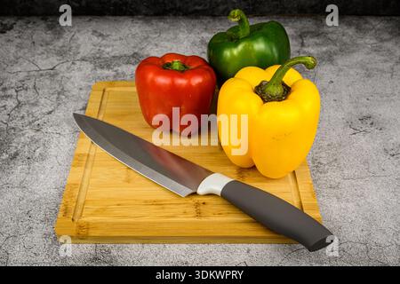 Rote, grüne und gelbe Paprika mit einem Kochmesser auf einem hölzernen Schneidebrett. Food Prep Stillleben auf grauem Hintergrund, gesundes Essen und Frühlingszwiebel Stockfoto