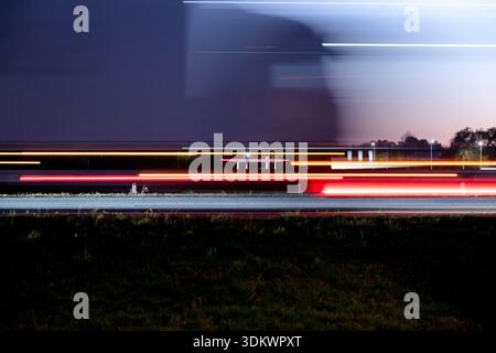 Verschwommene Lichtspuren eines Lastwagens und Verkehrsgeschwindigkeit an einer Tankstelle in der Abenddämmerung vorbei. Horizontale Ansicht mit langer Belichtung und Kopierraum für Business-Editorial. Stockfoto