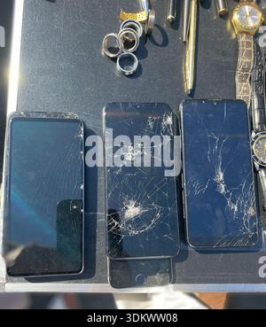 Drei beschädigte Smartphones mit gerissenen Bildschirmen werden auf einer reflektierenden Oberfläche platziert, begleitet von Ringen, einem Stift und einer Armbanduhr in hellem Licht Stockfoto