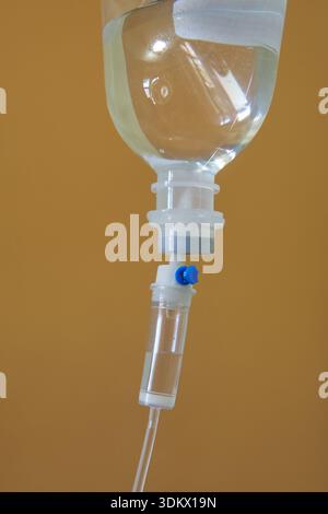 Intravenöse Tropfflasche isolierter brauner Hintergrund, klarer Plastikbehälter mit Kochsalzlösung und blauem Ventil auf medizinischem Schlauch, Nahaufnahme Seitenansicht Stockfoto