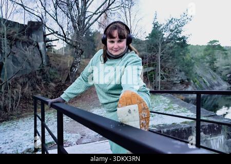 Die weibliche Athletin dehnt die Beine am Geländer an einem ruhigen Ort im Freien und trägt warme grüne Kleidung, die die Fitness und das Engagement für Heilung betont Stockfoto