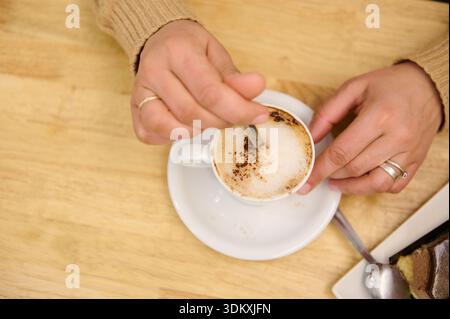 Nahaufnahme von Händen, die einen schaumigen Latte in einer weißen Tasse rühren, Ringe sichtbar, auf einem warmen Holztisch mit einem Dessertteller in der Nähe. Gemütliches, intimes Café Stockfoto